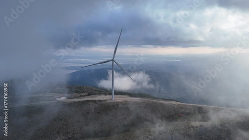 An aerial view of a wind turbine on a mountain top wind farm in between a  break in the morning misty cloud.