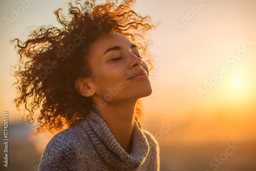 Young woman basking in golden hour sunlight, embracing peace and serenity with eyes closed, wind gently tousling her curly hair.