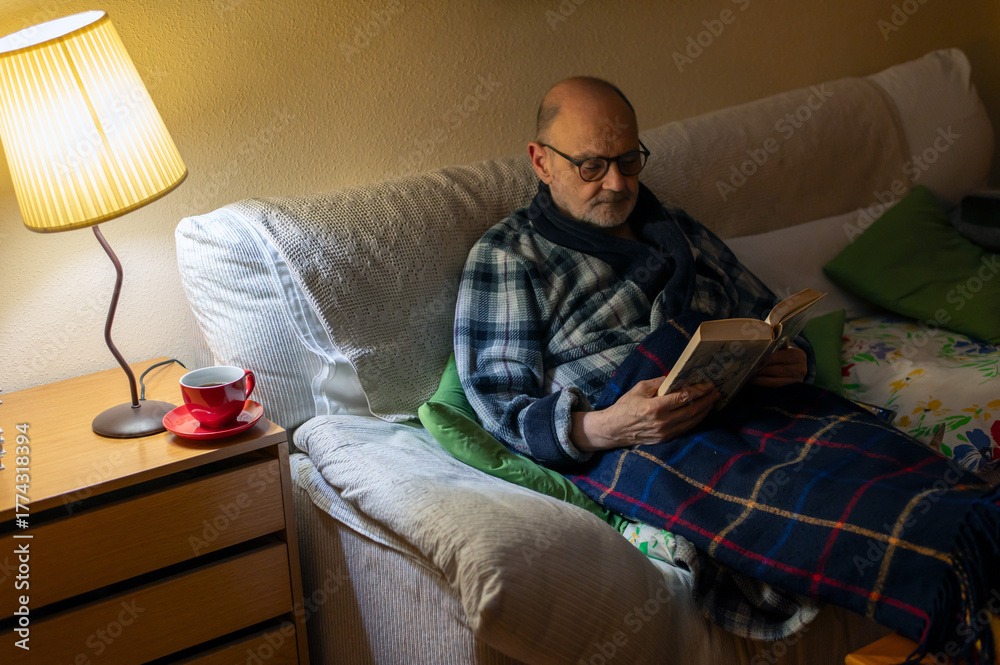 Fototapeta premium Older Man Reading Book on Sofa With Blanket, Lamp, and Cozy Home Vibe