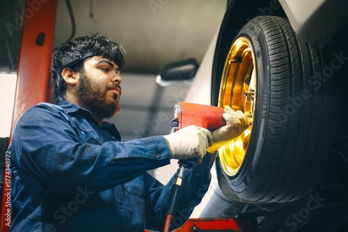 A mechanic in a blue uniform using a power tool on a golden car wheel