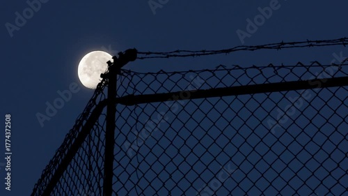 Time Lapse. Moon Behind Barbed Wire Fence At Night Over Chain Link Silhouette 
