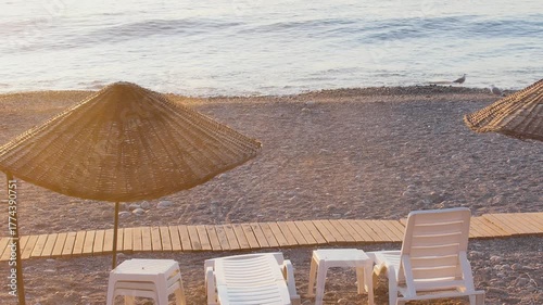 A picturesque, peaceful pebble beach scene at sunrise or sunset, featuring two thatched straw umbrellas and white plastic lounge chairs.