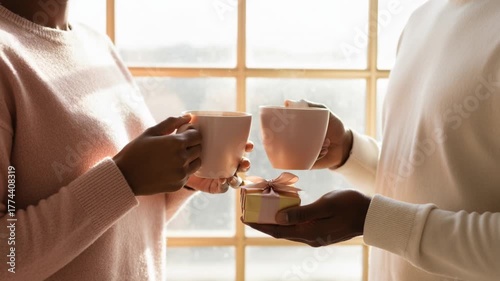 A black couple holding coffee mugs and a small gift. Close-up on an intimate moment by a sunlit window. Romance and celebration concept