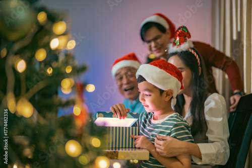 Excited Asian boy opening a Christmas gift with family cheering beside him, creating a heartwarming moment filled with love and festive happiness.