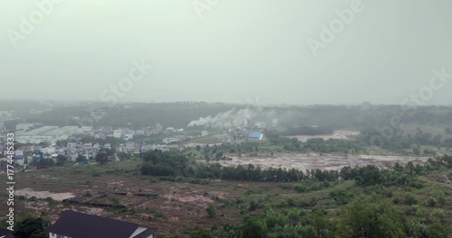 On a rainy, hazy day, smoke rises over a developing landscape featuring a large, muddy construction site in the foreground.