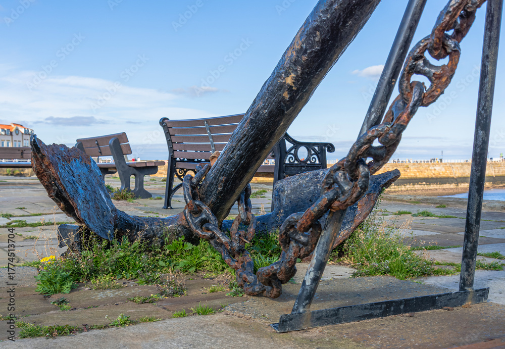Fototapeta premium An ancient weathered anchor is on a quay with weeds growing around it.
