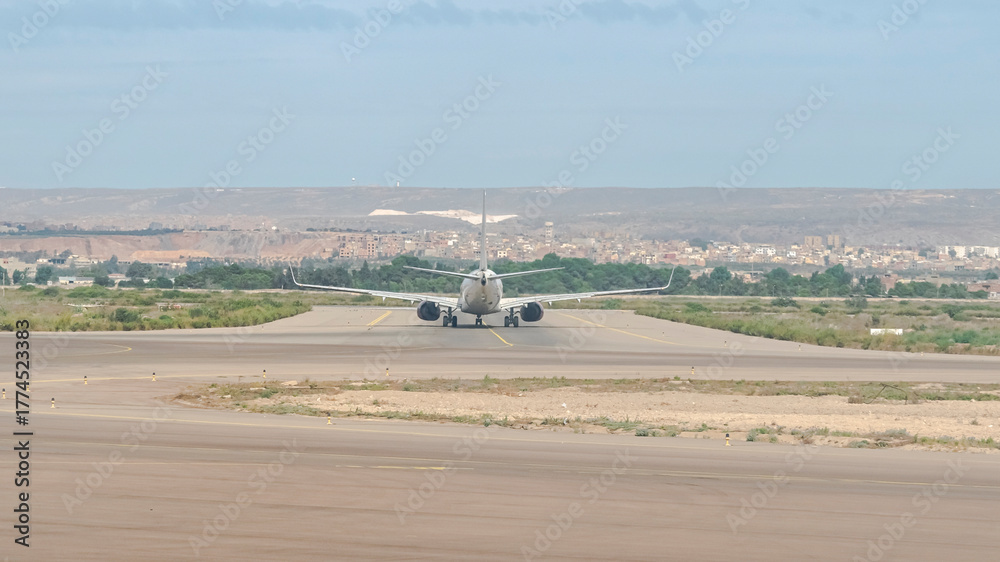 Fototapeta premium Rear view of a commercial airplane taxiing on the runway preparing for takeoff, with forest trees, old city buildings and a distant mountain under a clear blue sky.