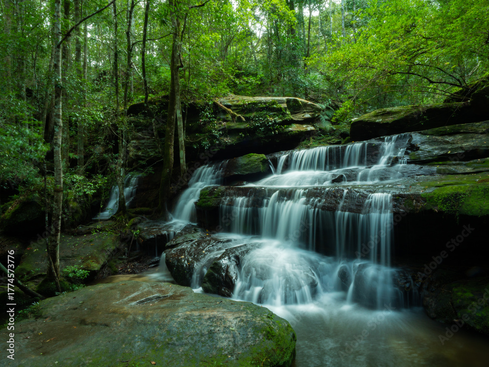 Fototapeta premium The waterfall looks smooth gentle, multi-tiered, beautiful flowing over mossy rocks in the lush green forest behind, dense lush, with tall trees dense bushes, Tourist attractions in Thailand