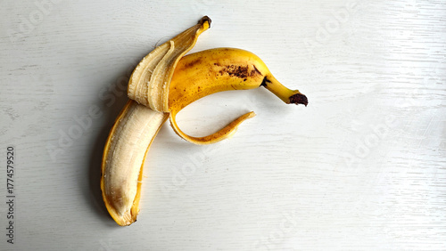 Half-peeled banana with skin lying on white wooden surface, top view. Banana has some brown spots, indicating it ripe. Tropical snack rich in nutrients. Healthy eating, diet
