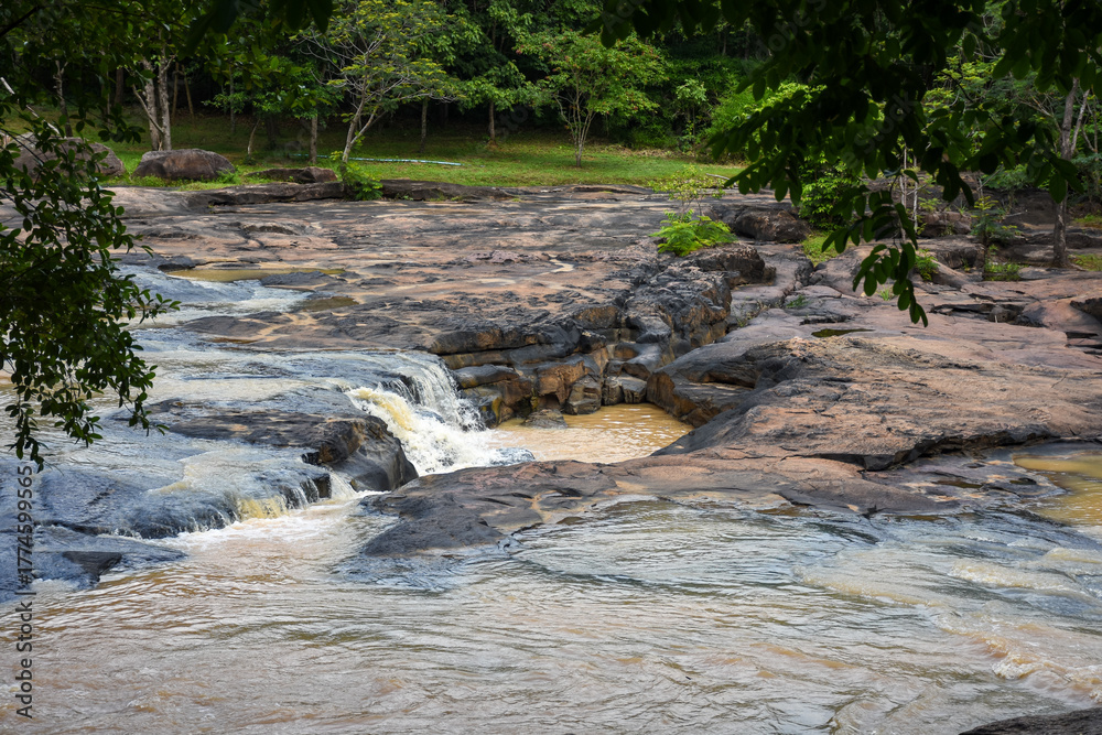 Fototapeta premium A fast-flowing, muddy river over dark rocks in a forest.