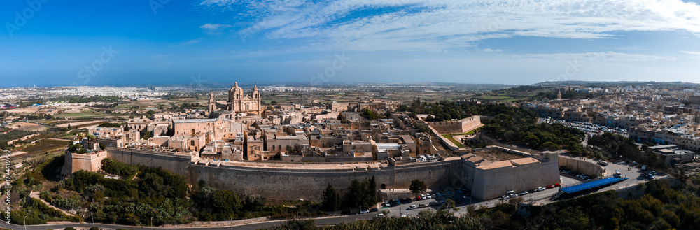 Fototapeta premium Aerial panorama of Mdina, Malta, with St. Pauls Cathedral, thick limestone bastion walls, and rural fields under crisp light, contrasting with modern suburbs beyond.
