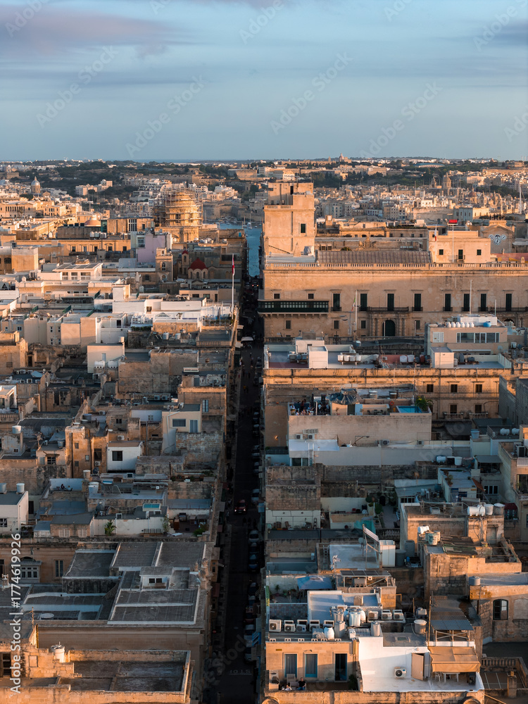Obraz premium Aerial view of Valletta, Malta, at sunset. A straight street cuts through limestone blocks toward a domed church and fortified palazzi, with deep shadows and warm light.