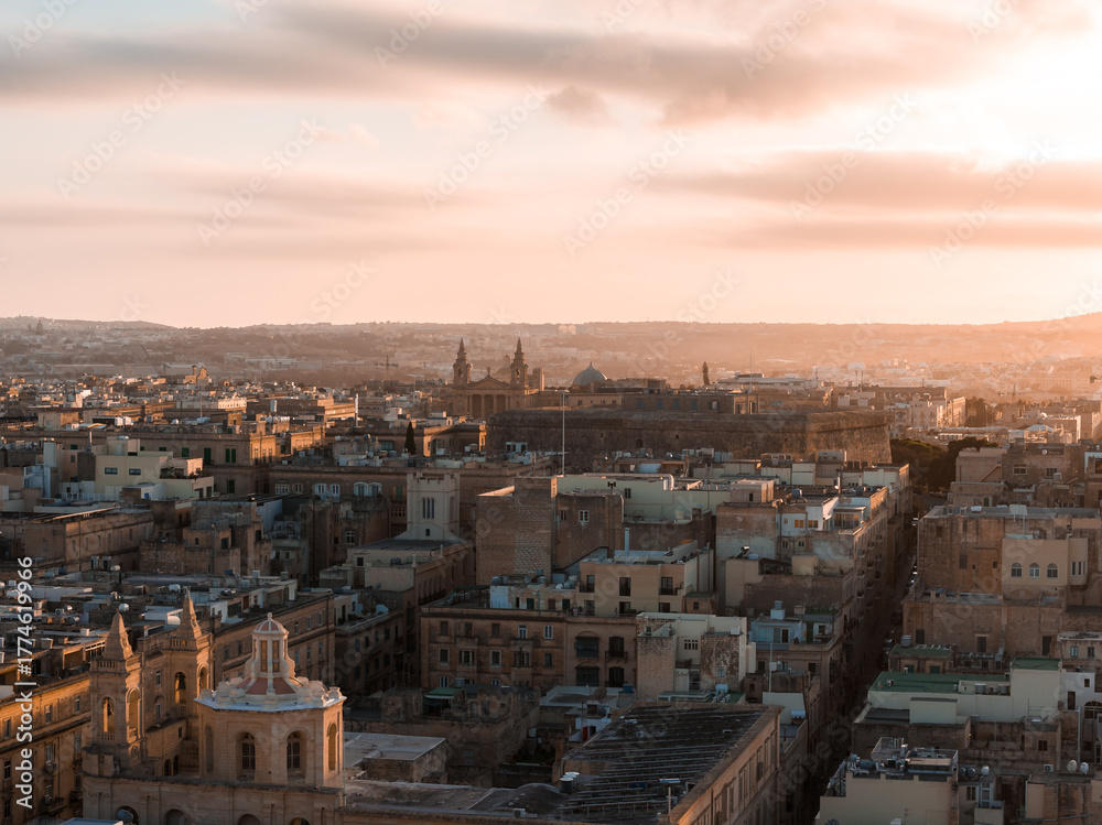 Obraz premium Aerial view of Valletta, Malta at golden hour, warm light on limestone blocks, church domes and twin spires, narrow streets lead to the harbor, long shadows add depth.