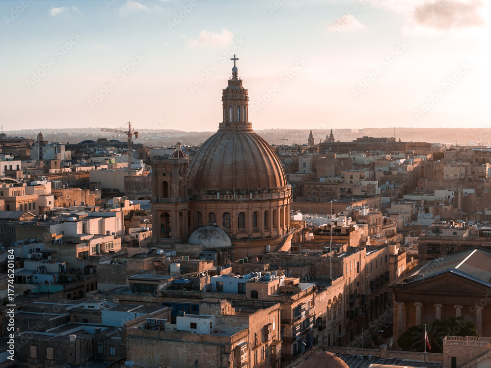 Fototapeta premium Aerial view shows Valletta, Malta at golden hour, the basilica dome and lantern above limestone blocks, long shadows across Baroque streets near Grand Harbour.