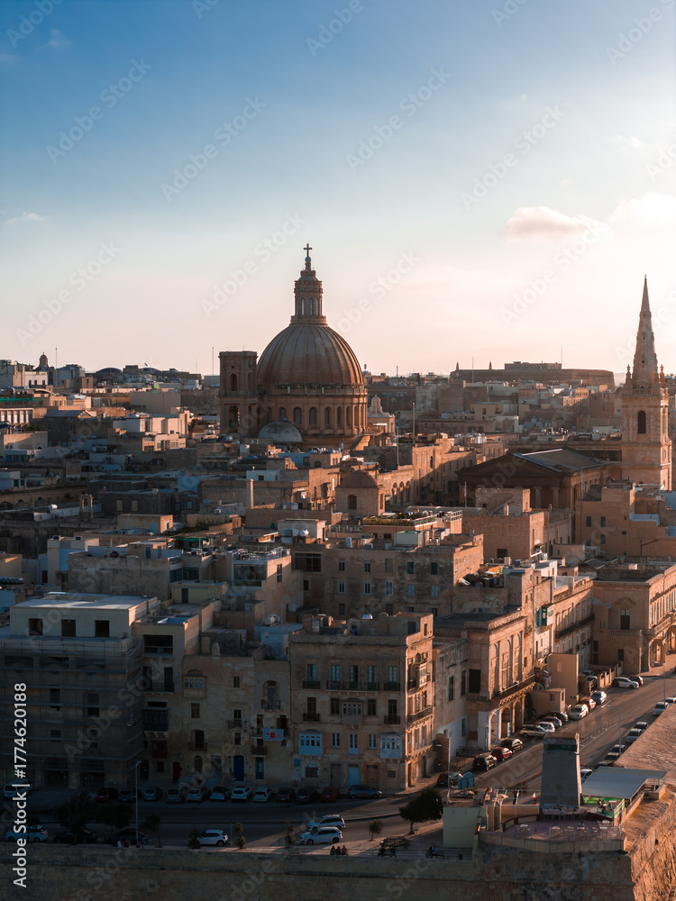 Naklejka premium Aerial view of Valletta, Malta at sunset shows the Basilica dome and St. Pauls Pro Cathedral spire over limestone blocks, warm golden light, long shadows, and a waterfront road.