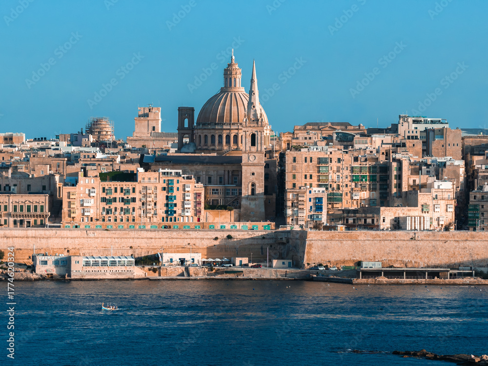 Naklejka premium Valletta, Malta, seen across Grand Harbour, shows the basilica dome, cathedral spire, stone walls, and a small boat in warm late day light with clear blue color.