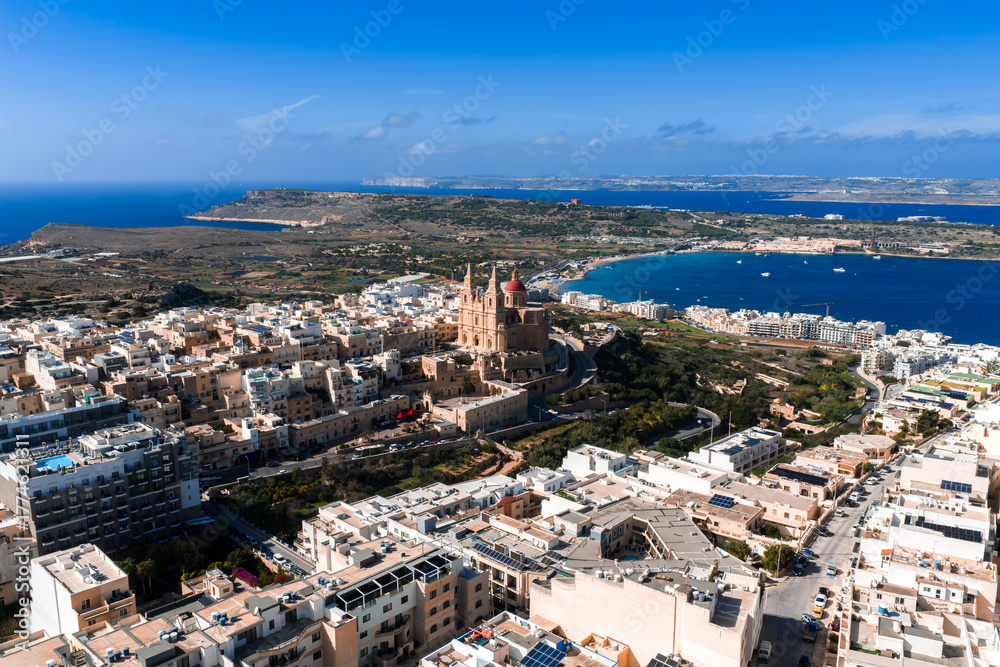 Fototapeta premium Aerial panorama of Mellieha, Malta shows Mellieha Parish Church above limestone buildings, Mellieha Bay, and Comino and Gozo on the horizon in bright daylight.