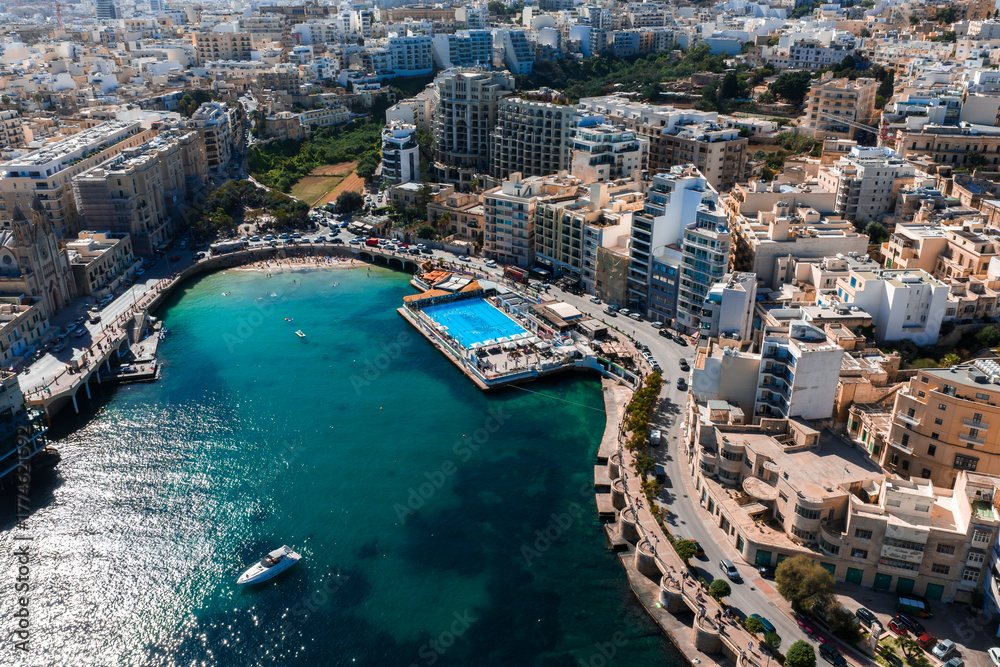 Fototapeta premium Aerial view of Sliema, Malta shows a turquoise inlet, a seaside lido with a large rectangular pool on a pier, curving apartments, a promenade, light traffic, and a drifting boat.