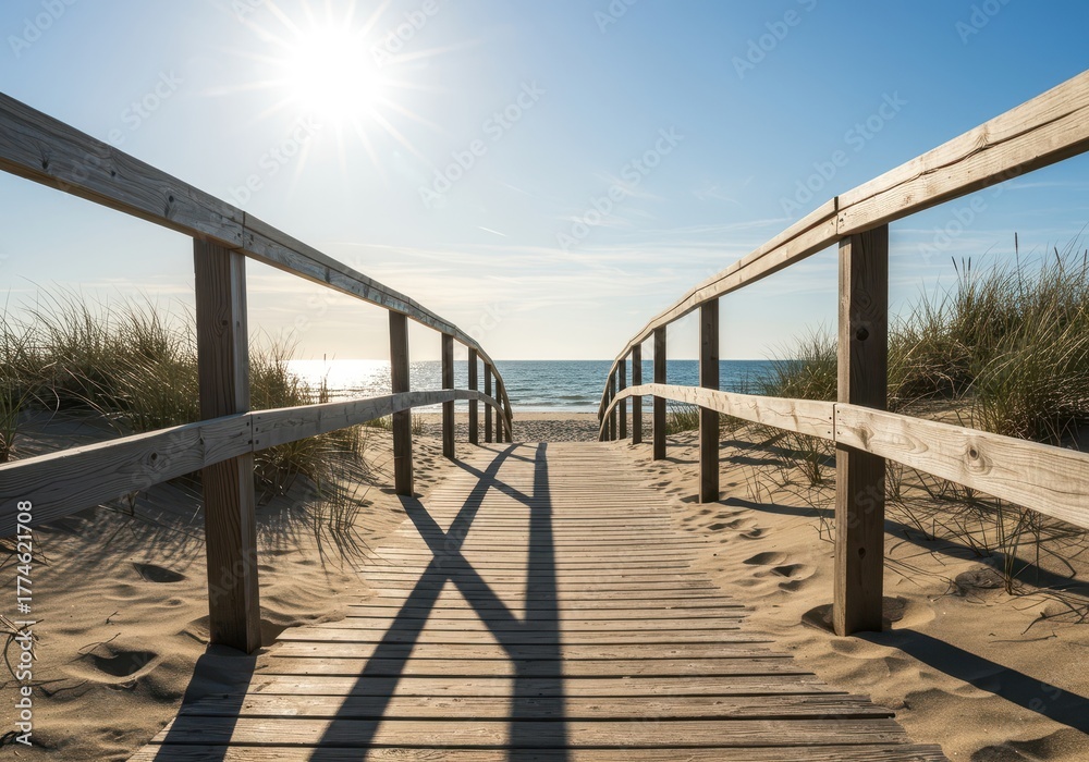 Fototapeta premium Coastal wooden walkway extending over sandy terrain toward the sea, illuminated by bright sunlight, suggesting seaside recreation, platform, stroll, escape
