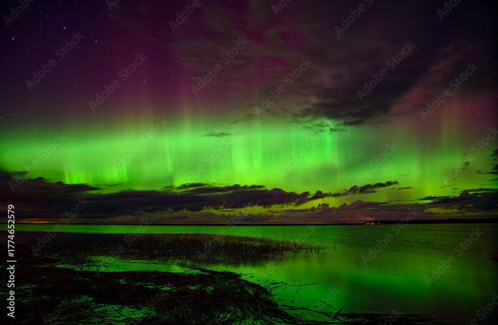 Fototapeta premium Vibrant aurora borealis in green and purple hues illuminating the night sky over a calm lake with reflections and clouds.