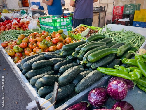 A colorful assortment of fresh organic vegetables displayed at an outdoor farmers market. Vibrant tomatoes, cucumbers, zucchini, eggplants, and green peppers fill the stall