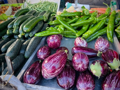 A colorful assortment of fresh organic vegetables displayed at an outdoor farmers market. Vibrant tomatoes, cucumbers, zucchini, eggplants, and green peppers fill the stall