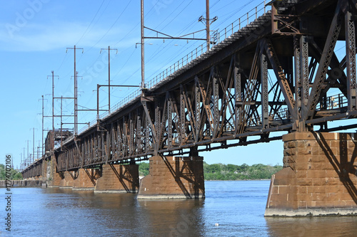 Industrial Truss Railroad Bridge Spanning a River Under a Clear Blue Sky, Highlighting the Steel Architecture, Utility Lines, and Surrounding Landscape on a Sunny Day