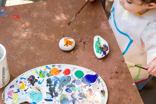 A young child is sitting at a table, painting stones with bright colors while surrounded by art supplies. The scene captures the joy of creative expression in nature.