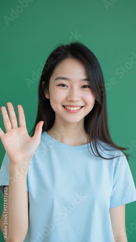Beautiful Asian girl in a light blue casual T-shirt smiling warmly and waving at the camera, standing against a solid green background, soft daylight, realistic style, friendly atmosphere.