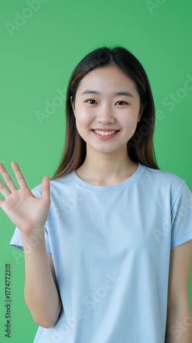 Beautiful Asian girl in a light blue casual T-shirt smiling warmly and waving at the camera, standing against a solid green background, soft daylight, realistic style, friendly atmosphere.