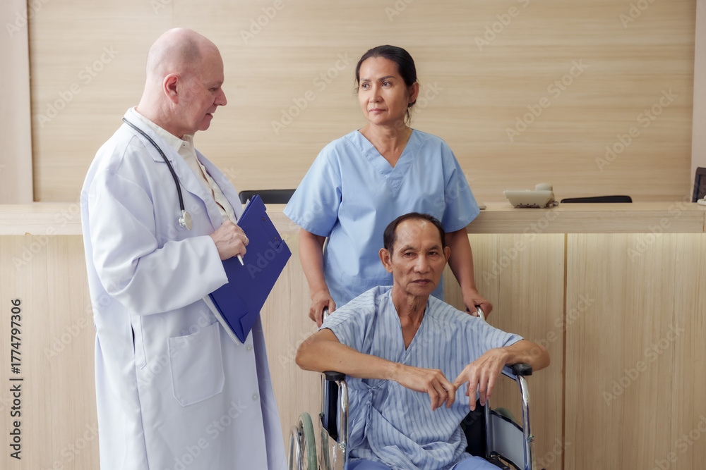 Obraz premium Senior asian male patient sitting in wheelchair receiving medical consultation from senior caucasian male doctor with female nurse standing beside at hospital reception area