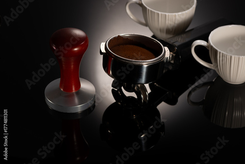 Espresso machine portafilter handle with compacted coffee and wooden tamper over gradient black background with reflection.