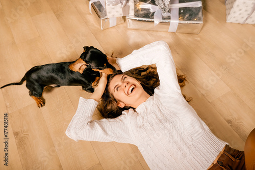 One cheerful girl in a white knitted sweater and brown pants. A curly woman lies on the floor with a black dachshund dog