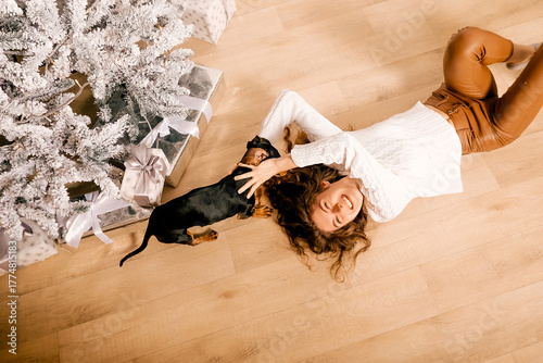 One cheerful girl in a white knitted sweater and brown pants. A curly woman lies on the floor with a black dachshund dog