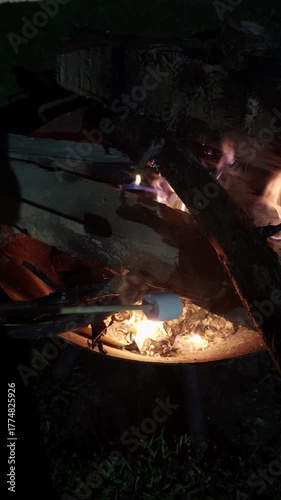 Stick with a marshmallow held near an outdoor campfire with glowing flames and burning wood. Warm evening camping scene