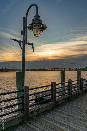 A peaceful sunset illuminates the Wilmington, North Carolina Riverwalk, where a vintage lamppost stands over a wooden boardwalk beside the calm Cape Fear River.