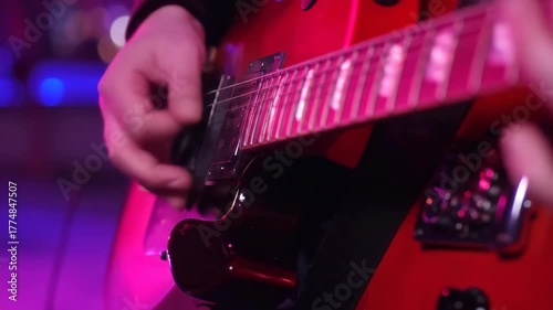 Close-up of hands playing a red electric guitar under vibrant pink and purple stage lights, dynamic