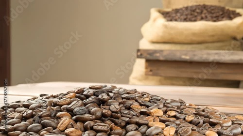 A heap of whole coffee beans on a wooden surface, with blurred jute sacks filled with beans behind