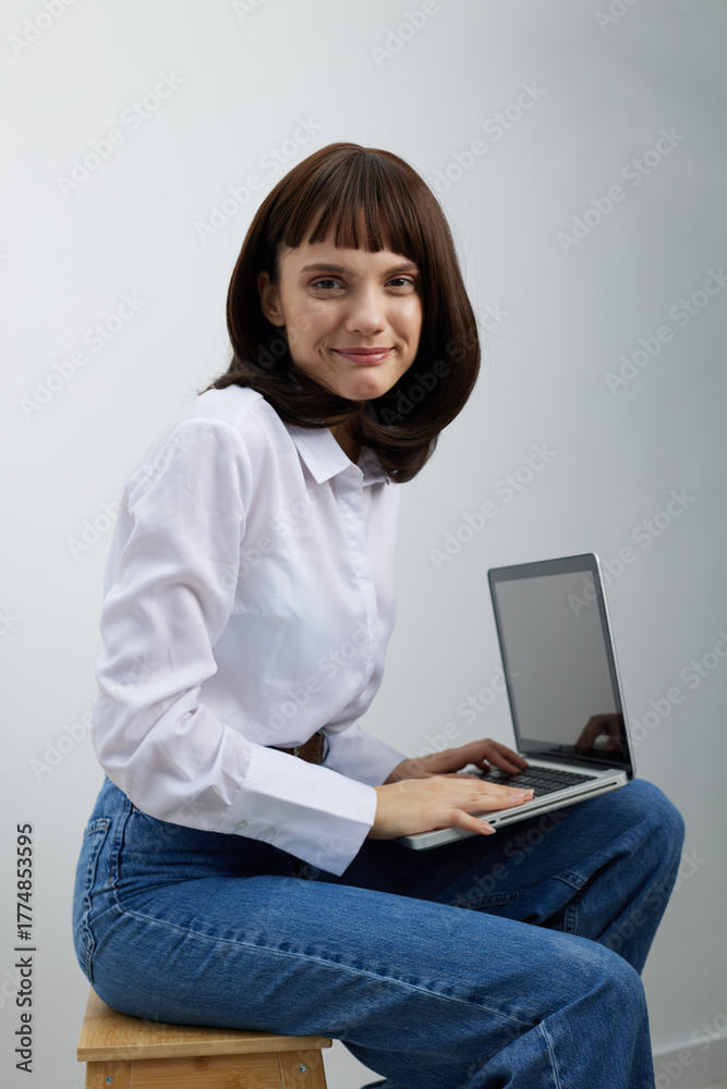 Naklejka premium A focused young woman sits on a wooden stool with a laptop in a bright, minimal space. She wears a white shirt and blue jeans, creating a casual yet productive mood for modern work, study, or