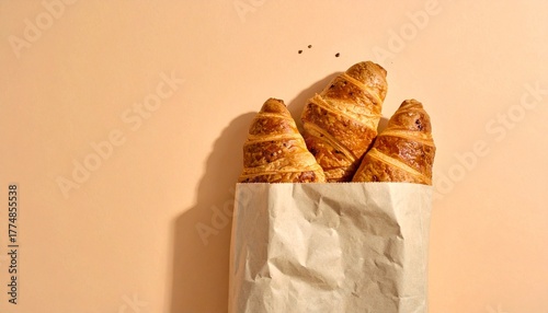 Three golden brown croissants peek out from a crumpled white paper bag, set against a soft peach background