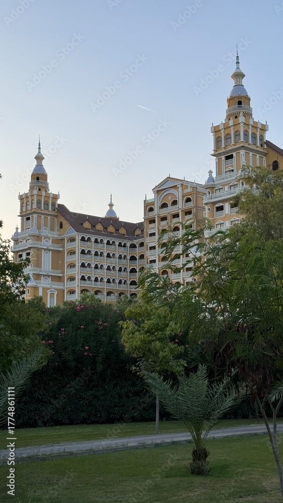 Fototapeta premium Majestic luxury hotel building featuring baroque-style yellow facades and ornate towers, partially obscured by green garden foliage and palm trees