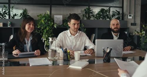 Over the shoulder Three office workers in business attire sit at a meeting table while one woman stands and greets group with a handshake in a modern office environment