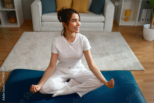 Smiling young Asian woman sits comfortably on a yoga mat in a relaxed meditation pose in her bright, modern living room at home.