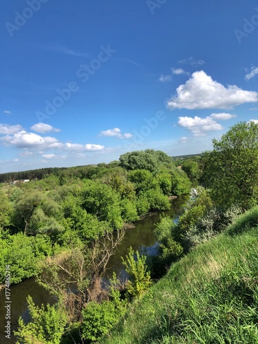 Idyllic Summer Landscape: River Flowing Under Dramatic Cloudy Sky. Emphasizes vibrant green summer foliage and the powerful contrast of the blue sky and cloudscape.