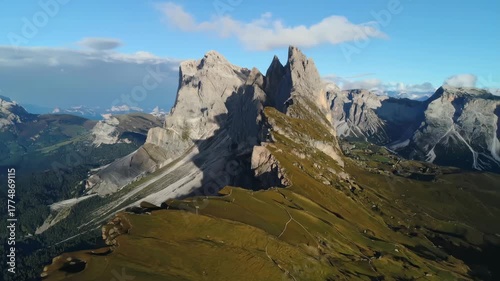 Dramatic jagged mountain peaks rise above lush green alpine meadows under a bright blue sky with scattered clouds