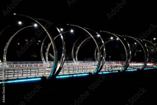 Modern illuminated pedestrian bridge with circular metal arches at night, contemporary architecture and urban design landmark with lights and reflections