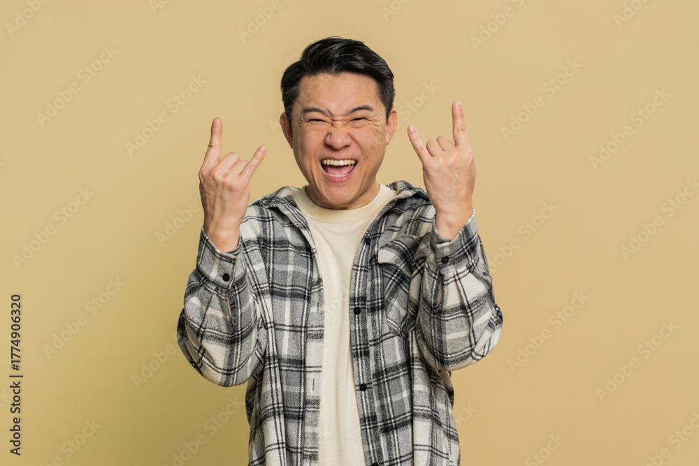 Naklejka premium Overjoyed happy Asian middle-aged man showing rock n roll gesture by hands cool sign shouting yeah with crazy expression, dancing, rejoicing victory win success. Mature Chinese guy on beige background