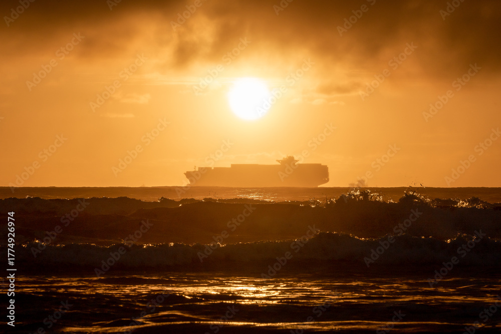Fototapeta premium Cargo ship sailing at sunset near the horizon with golden light reflecting on the water surface