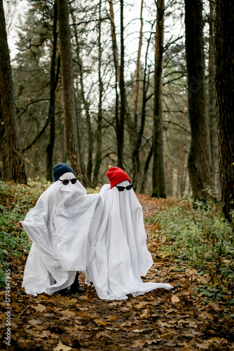 Children dressed as ghosts stand on a path in an autumn forest. The costumes are made of white fabric, and they wear black and red hats and sunglasses. This is a Halloween concept.