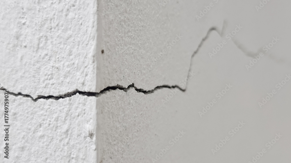 Obraz premium Close-up of a distinct diagonal crack in the corner of a rough white painted concrete or stucco wal, showing damage and decay.